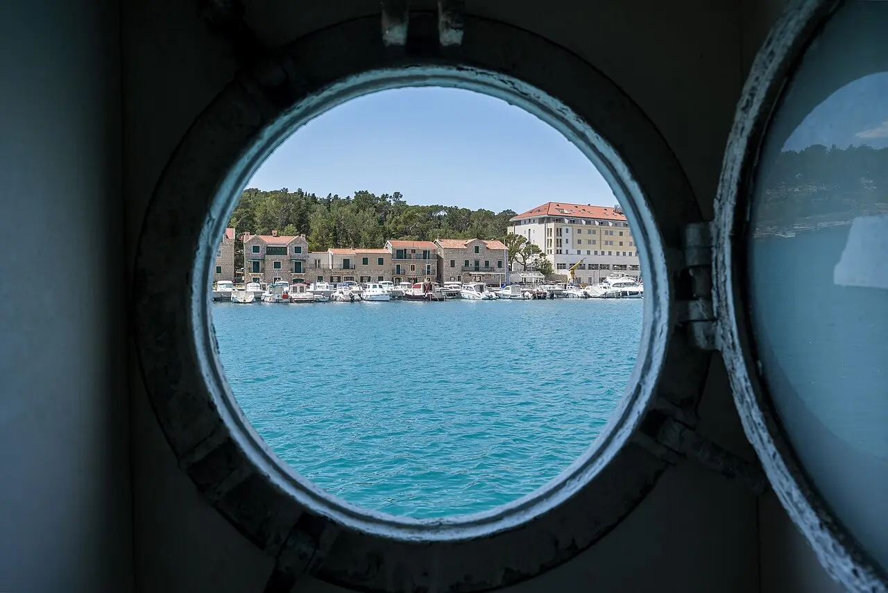 An island harbour of stone houses seen through the round porthole of a ferry