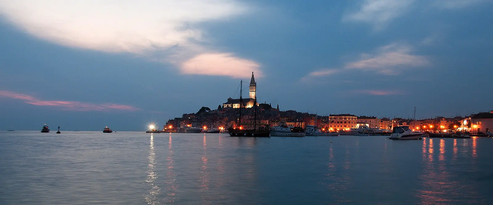 The skyline of Rovinj at dusk, its bell tower and old town silhouetted above the harbour lights and the sea