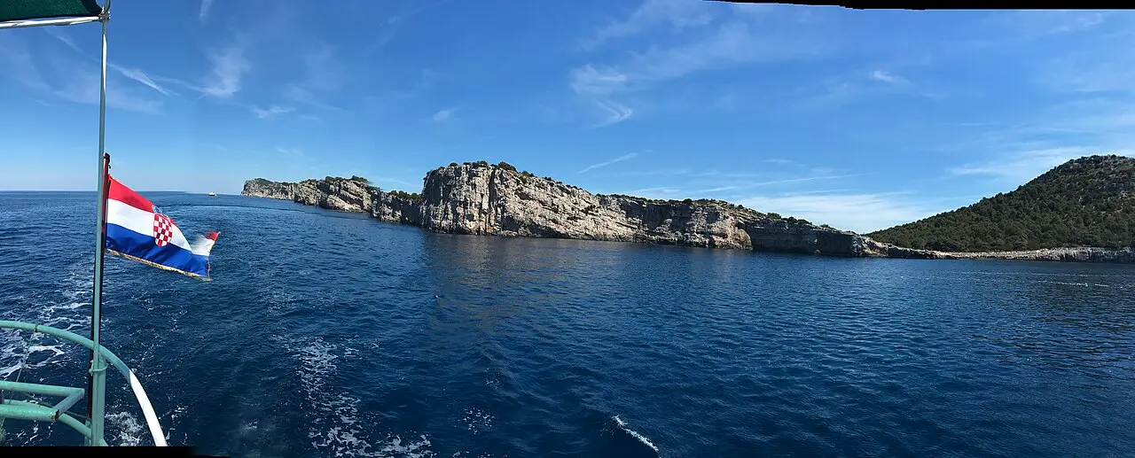 The steep white limestone cliffs of a Kornati island rising from deep blue sea, a Croatian flag on the stern of the tour boat