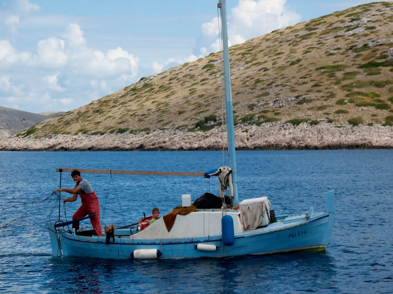 A fisherman in red waders casting a net from a traditional blue wooden boat registered in Murter, a bare Kornati island behind