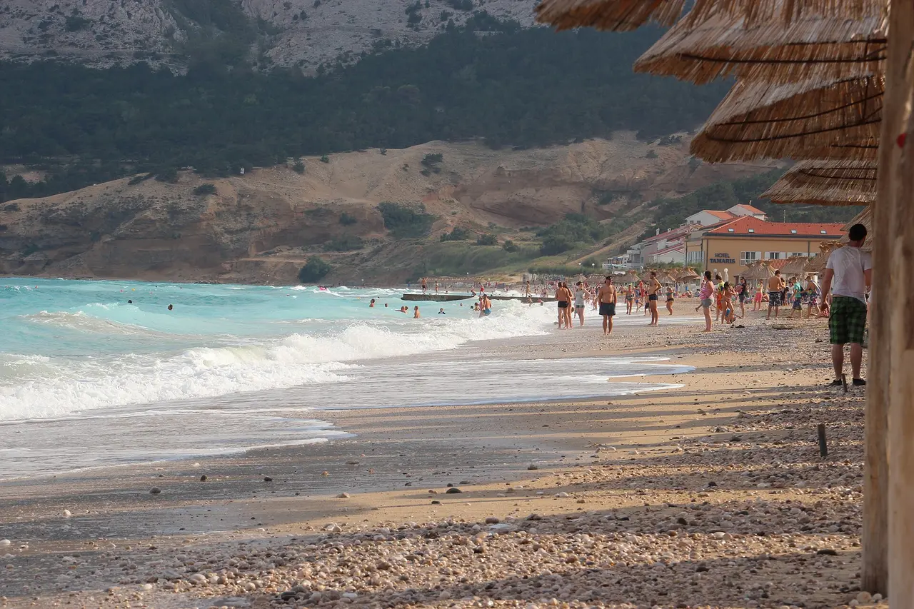 The long pebble beach of Vela plaza in Baska with bathers and straw parasols below bare hills