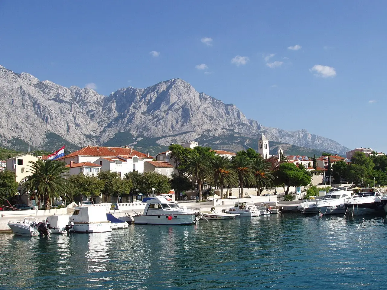 The harbour and palm-lined promenade of Baška Voda on the Makarska Riviera, with the grey Biokovo massif rising behind