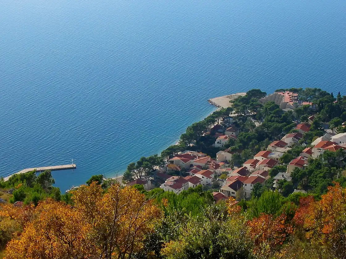 Aerial view of Brela on the Makarska Riviera, red-roofed houses among pines on a small peninsula with a pebble beach and a jetty