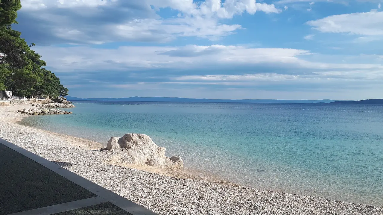 A white pebble beach at Brela on the Makarska Riviera, with clear turquoise water, a large white rock and pine trees along the shore