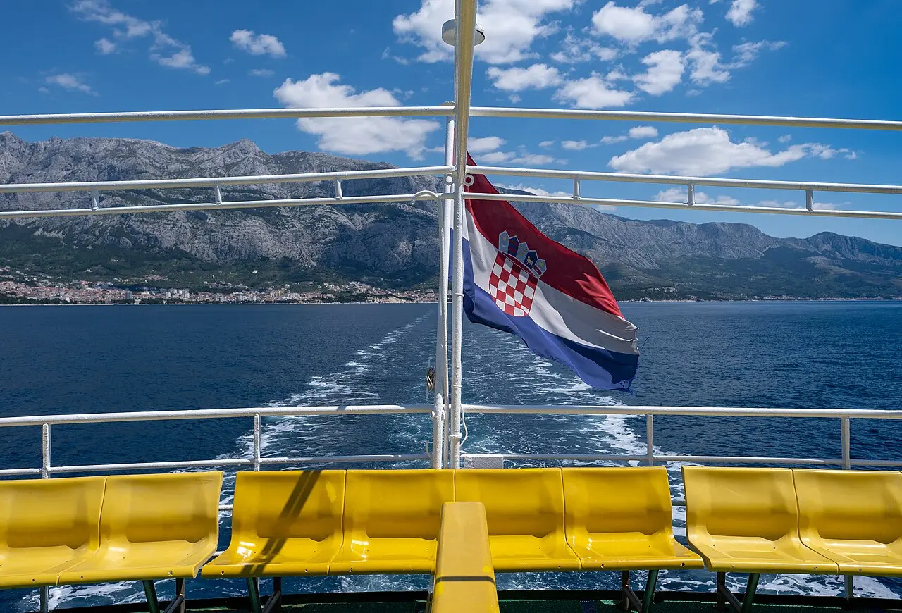 The Croatian flag flying from the stern of a ferry leaving Makarska, with the town strung along the coast and the Biokovo mountains behind