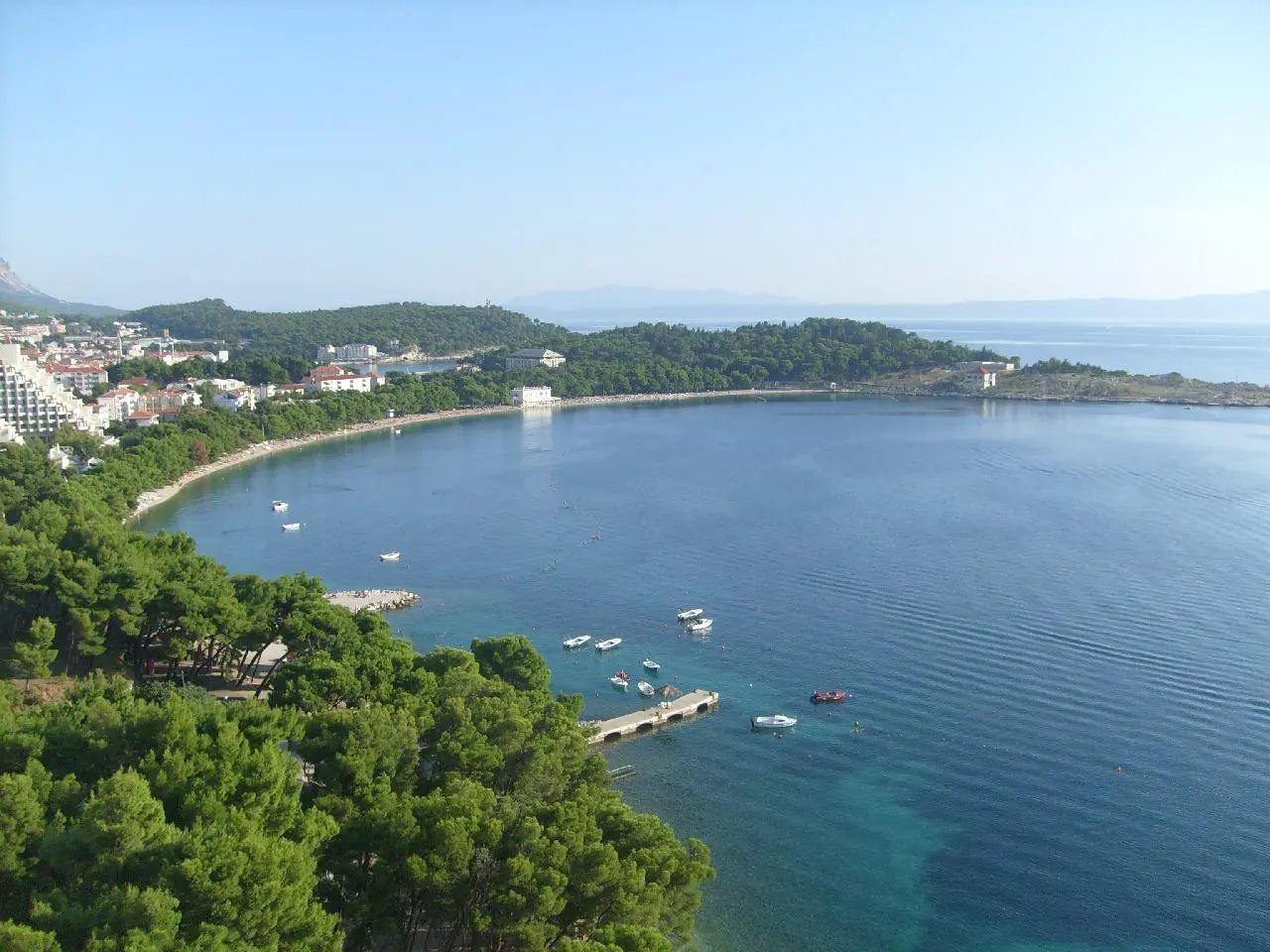 Makarska's curved bay and the wooded Sv. Petar peninsula seen from above, with boats moored and the town along the left shore