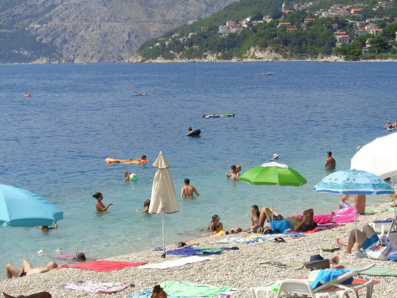 Sunbathers and umbrellas on the pebble Punta Rata beach at Brela, with clear water and a wooded point below Biokovo
