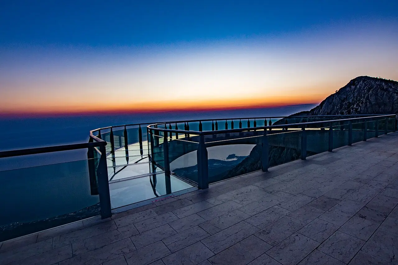 The semicircular glass Biokovo Skywalk platform jutting over the cliff at dusk, with the sea and a rocky peak beyond