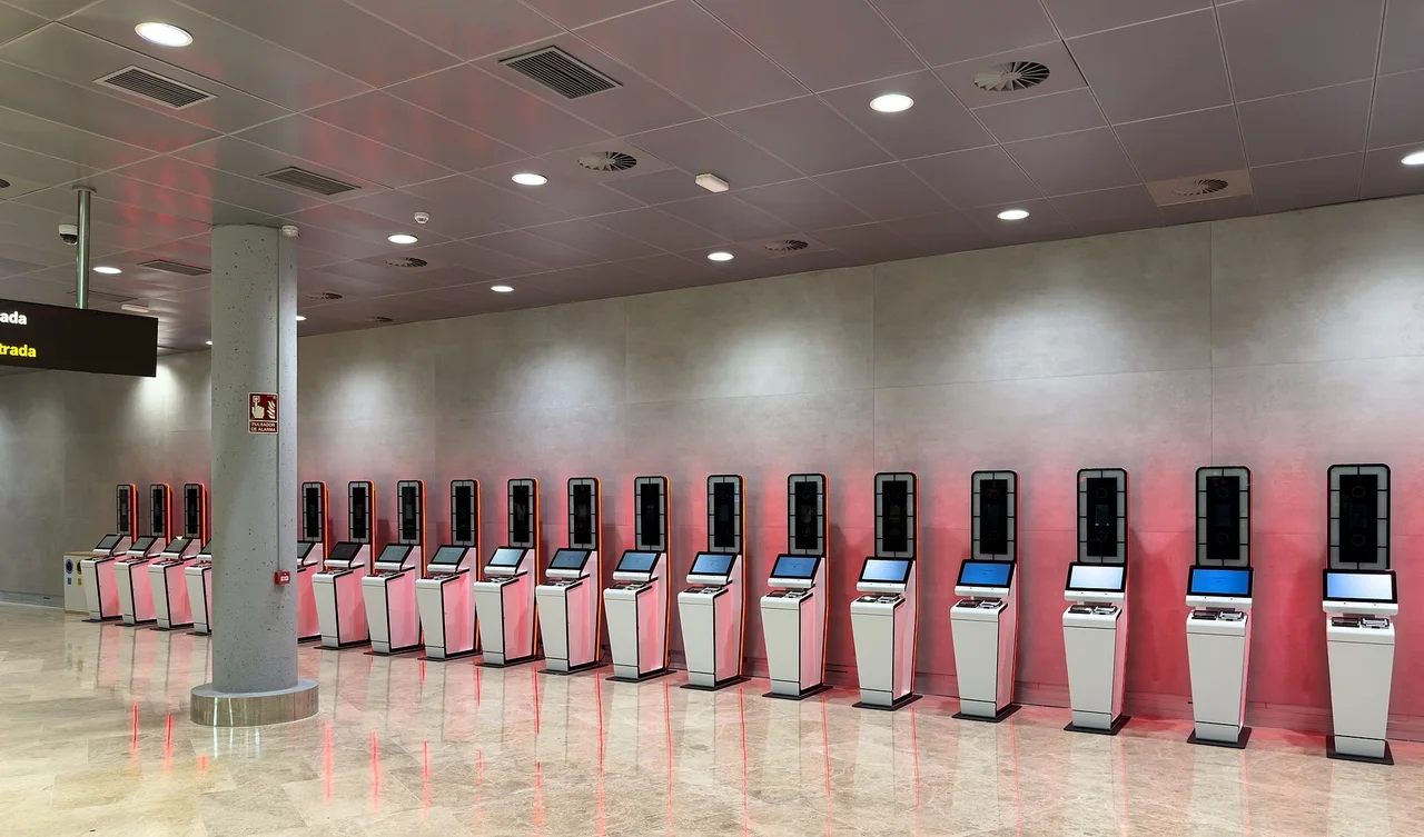A long row of white self-service passport kiosks with face-camera screens along a grey wall in an airport arrivals hall