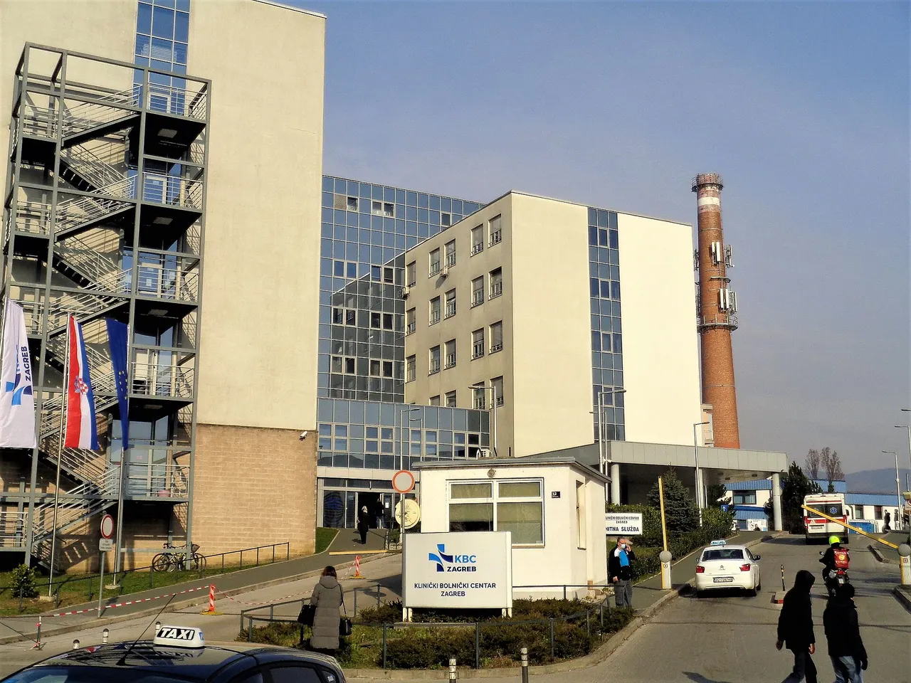 The entrance of the University Hospital Centre Zagreb at Rebro, with a KBC Zagreb sign, Croatian and EU flags, an ambulance and a taxi