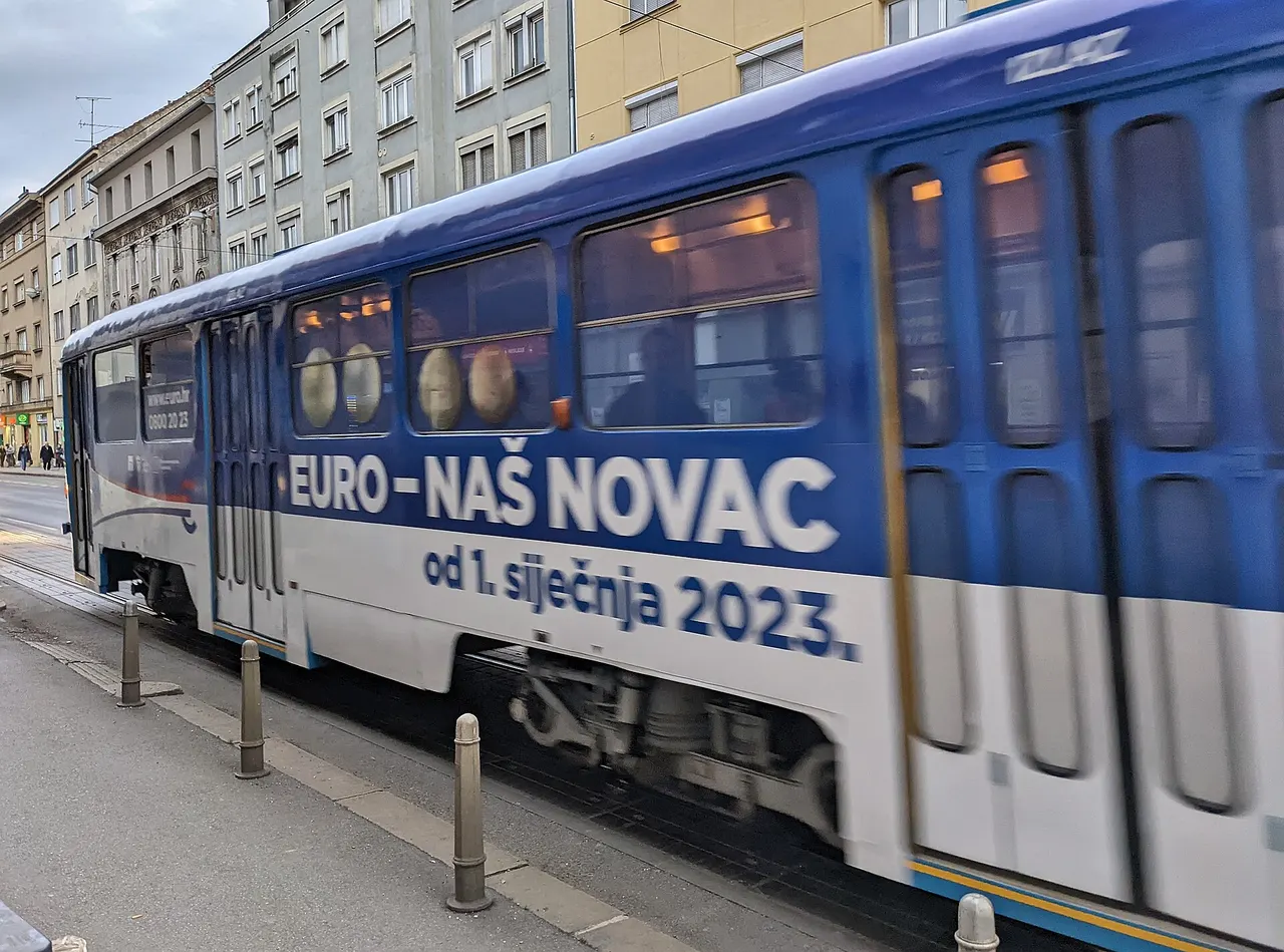 A blue and white Zagreb tram in motion, with the slogan Euro - naš novac, from 1 January 2023, and euro coins printed along its windows