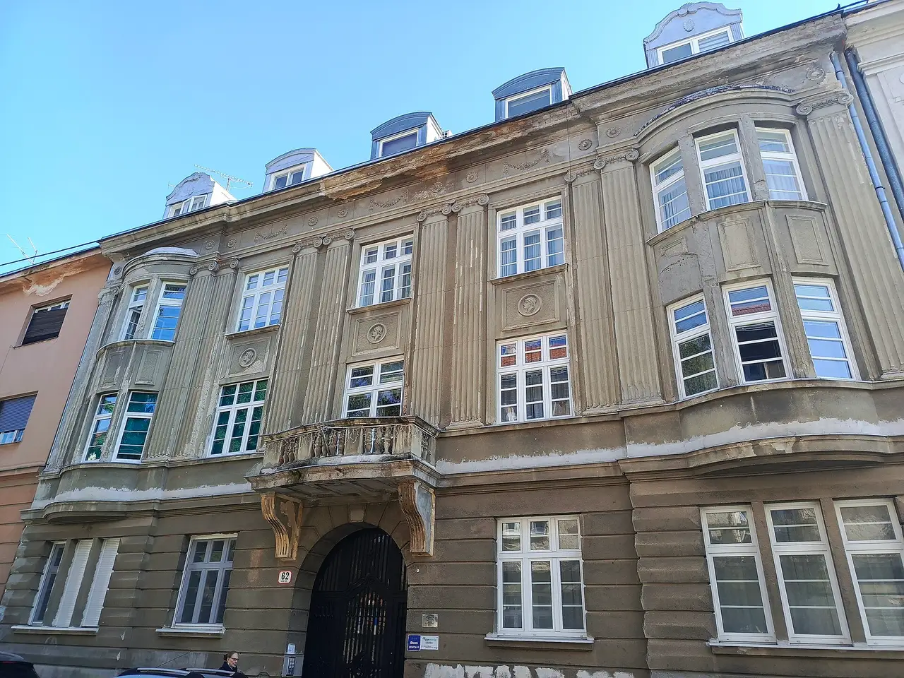 A three-storey apartment house in Zagreb with bay windows, dormers and an arched iron gate, the number 62 on the wall beside the entrance