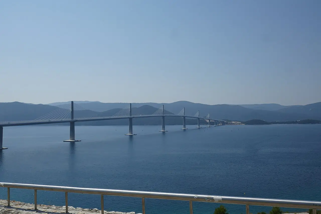 The full span of the Pelješac Bridge crossing the blue bay, seen from a coastal viewpoint with a railing in the foreground
