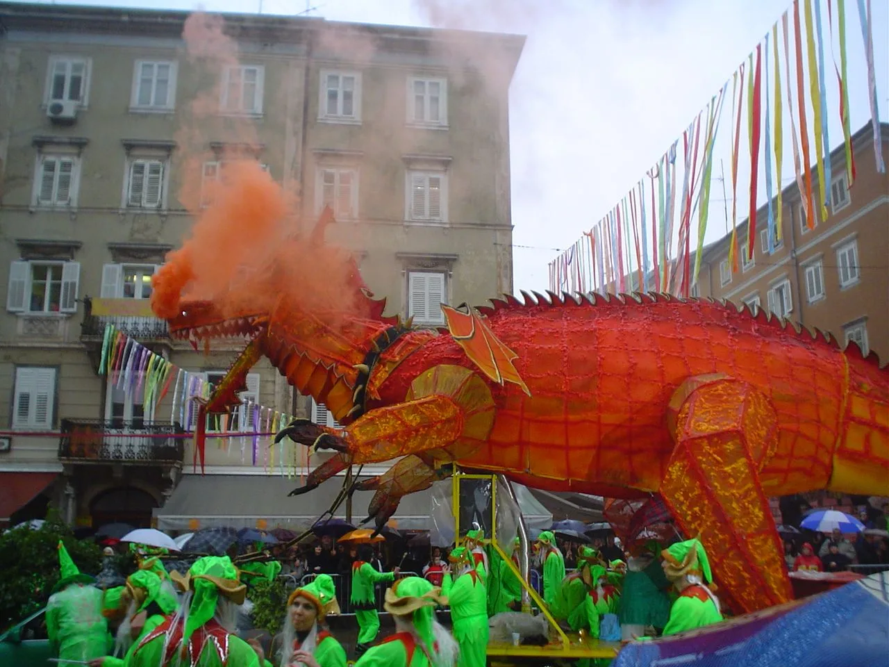 A huge orange dragon float breathing smoke above a carnival group in green costumes on a Rijeka street hung with streamers