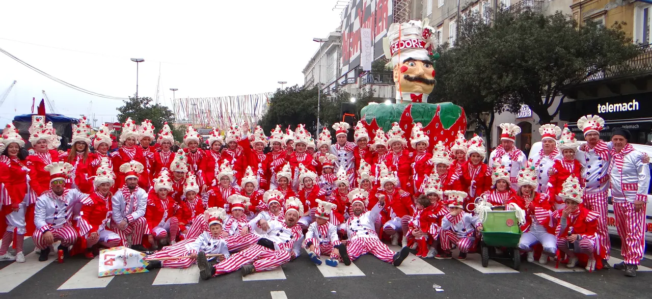 A large carnival group in red and white costumes posing on a Rijeka street in front of shop fronts and a giant figure on a float