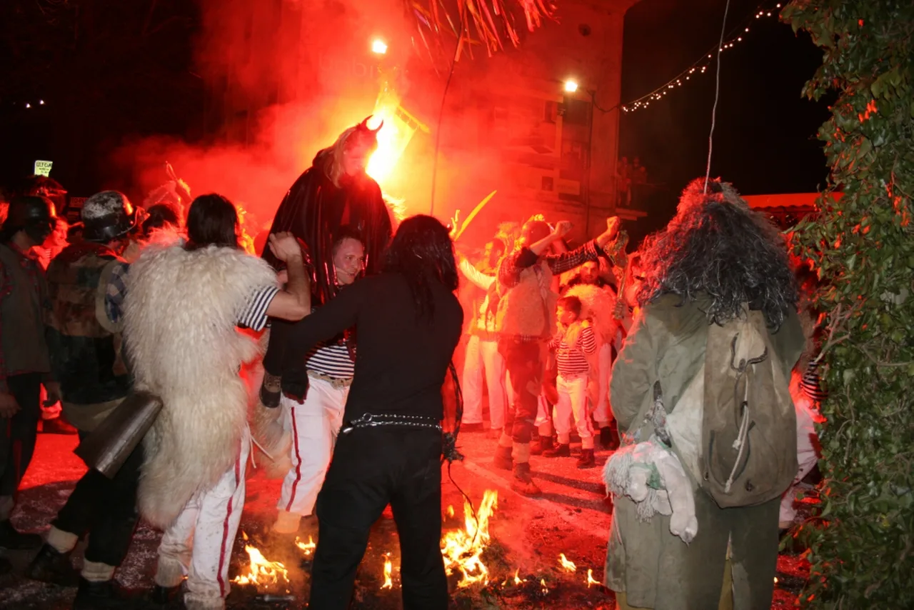Masked figures and bell ringers in sheepskins dancing around a bonfire in red flare light at night in Viškovo