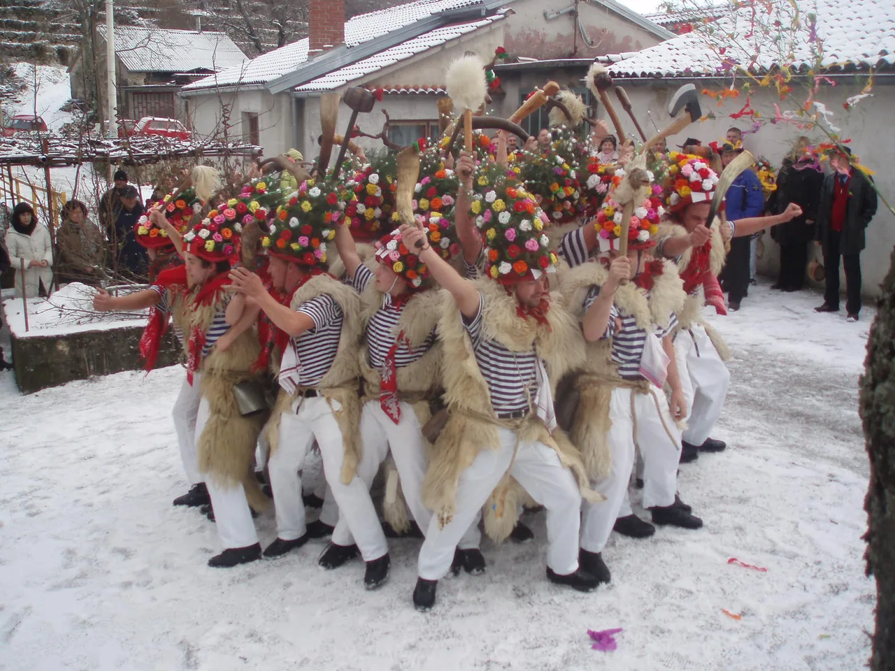 Bell ringers in sheepskins, striped shirts and tall hats covered in paper flowers forming a tight ring in a snowy village yard while neighbours watch