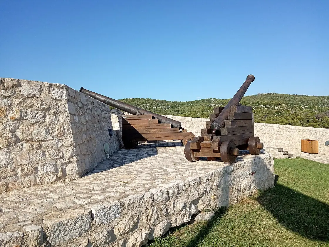 Two old wooden-carriage cannons on the stone ramparts of Barone Fortress above Šibenik, with wooded hills behind