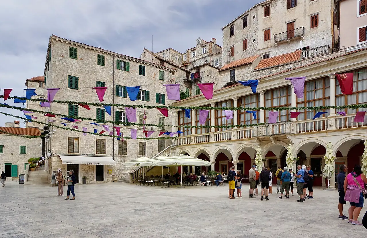 The main square of Šibenik with café tables, festive bunting and the arcaded stone town hall, stone houses climbing behind