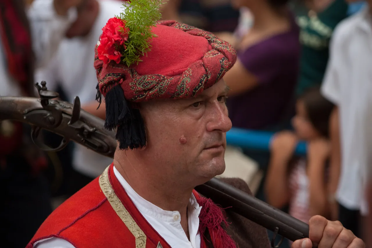 An Alka squire in a red cap decorated with a carnation and greenery, with an old long gun resting on his shoulder