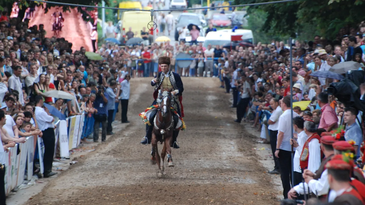 An alkar in a fur kalpak and dress uniform gallops down the Alka track in Sinj towards the camera, with the iron ring hanging from a rope above and crowds behind the barriers