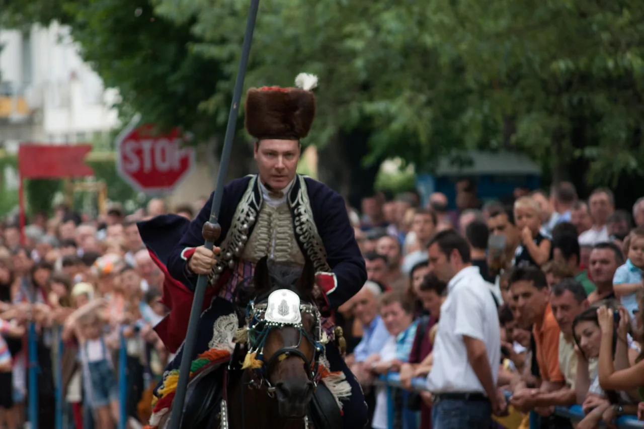 An alkar in a fur kalpak with a white plume and an embroidered dark blue jacket rides past the crowd holding his lance upright