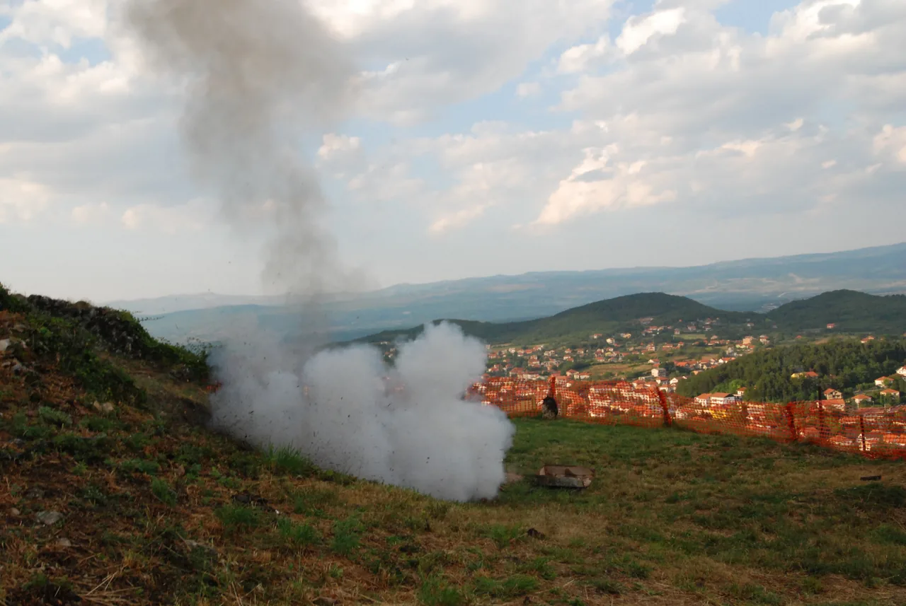 A cloud of white smoke rising from a ceremonial mortar on a grassy hillside above the red roofs of Sinj