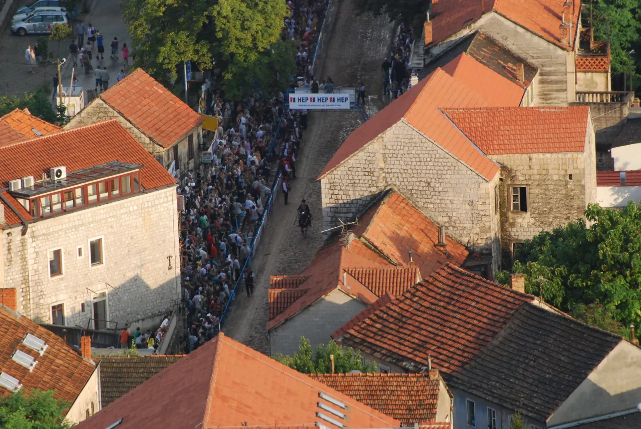 Aerial view of the Alka track running between stone houses with red tile roofs in Sinj, a rider on the track and a dense crowd along one side
