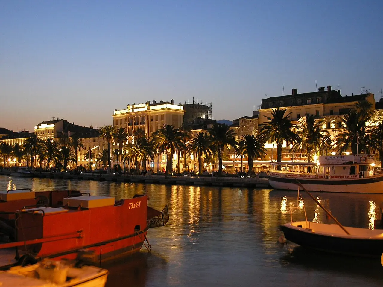 The Riva waterfront promenade in Split lit up at dusk, with palms, café terraces and boats reflected in the harbour