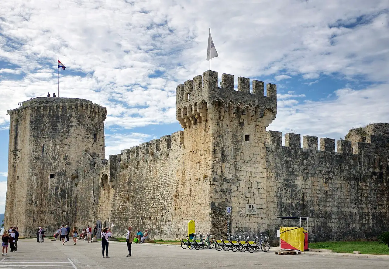 The round tower and crenellated stone walls of Kamerlengo fortress in Trogir, with visitors in front