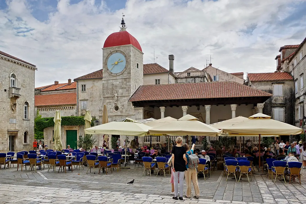 The main square of Trogir with the clock tower, the open town loggia and café tables under umbrellas