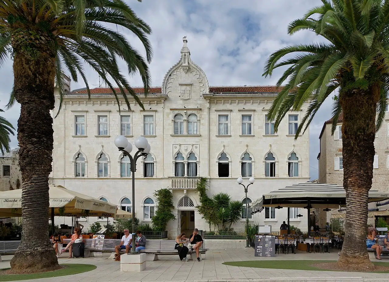 A grand pale Venetian-Gothic palace on the palm-lined Riva waterfront of Trogir, with benches and cafés