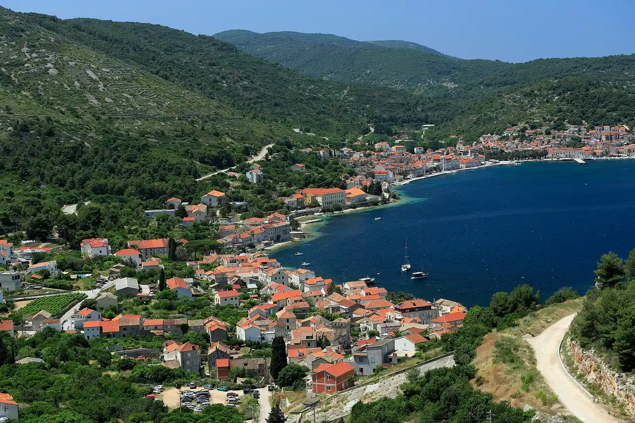 The waterfront of Vis town curving around its bay, with stone houses, palms and moored boats under a low green hill
