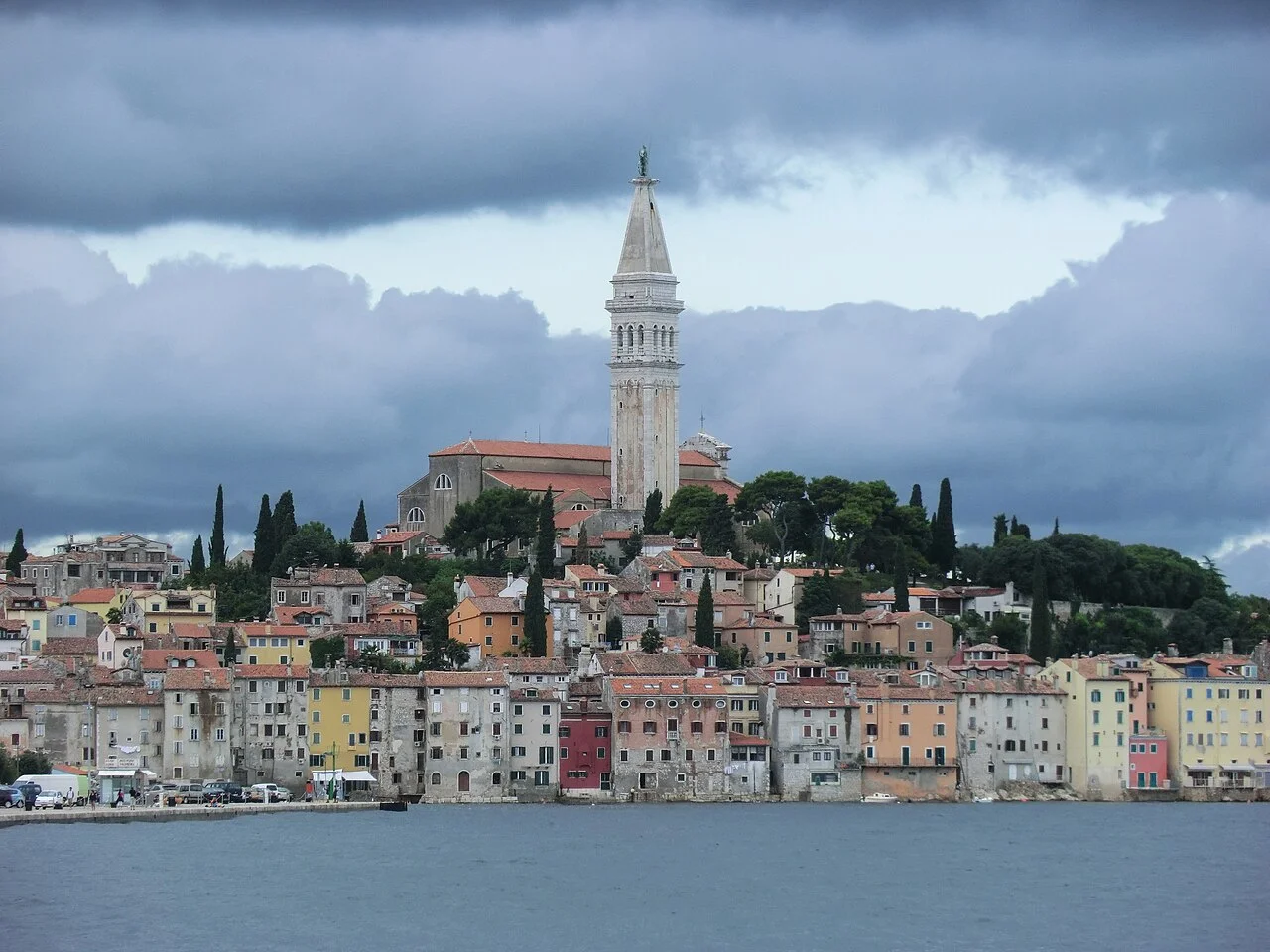 Colourful houses along the Rovinj old town waterfront with the St Euphemia bell tower rising above, seen from the water under a cloudy sky