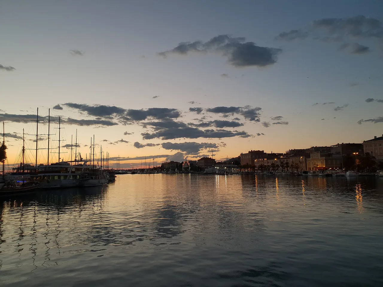 Boats moored along the Zadar waterfront at dusk, town lights and an orange sky reflected in the calm harbour