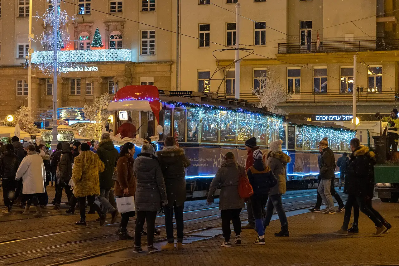 A vintage Zagreb tram covered in blue Christmas lights passing a crowd on Ban Jelacic Square in falling snow