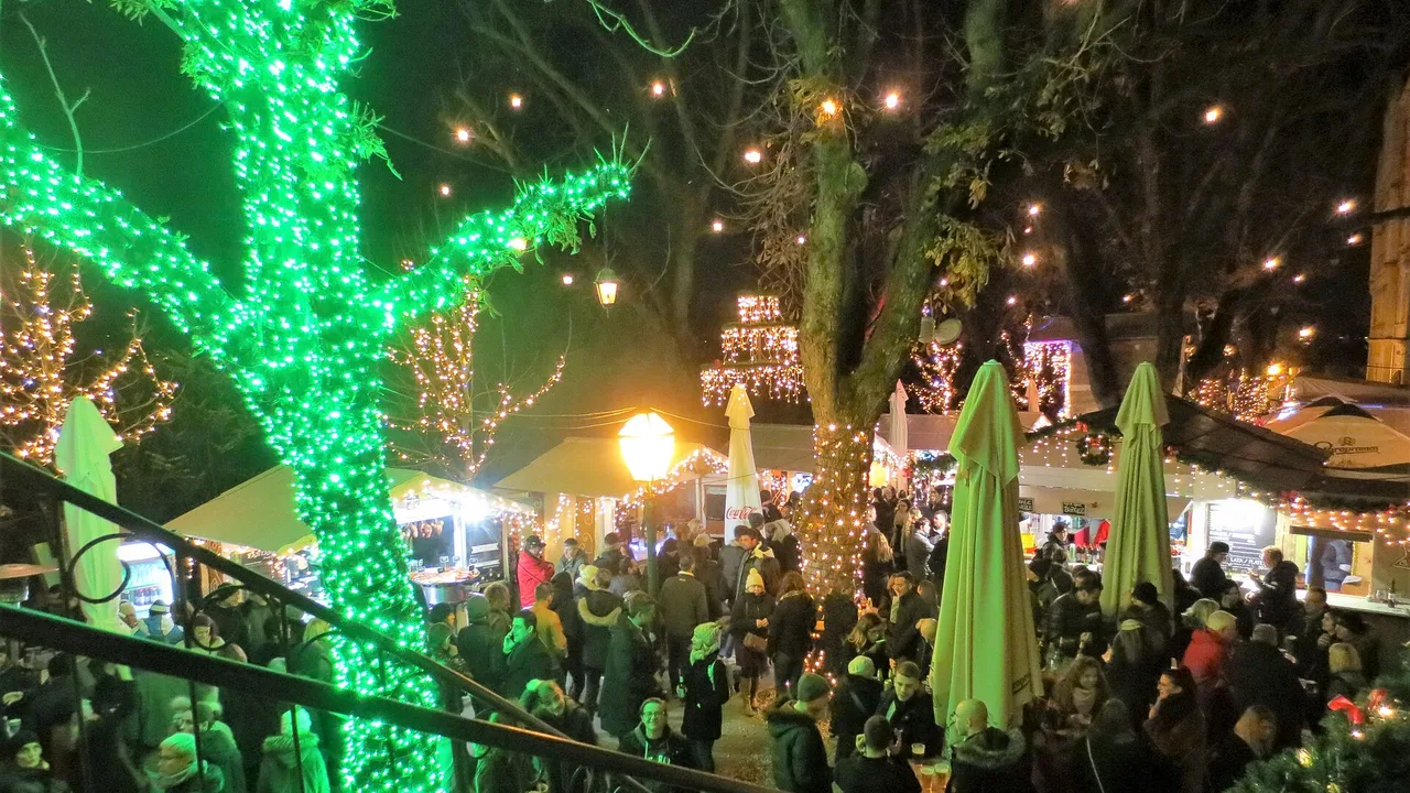 Crowds between food huts under plane trees wrapped in green and white lights at Advent in Zagreb