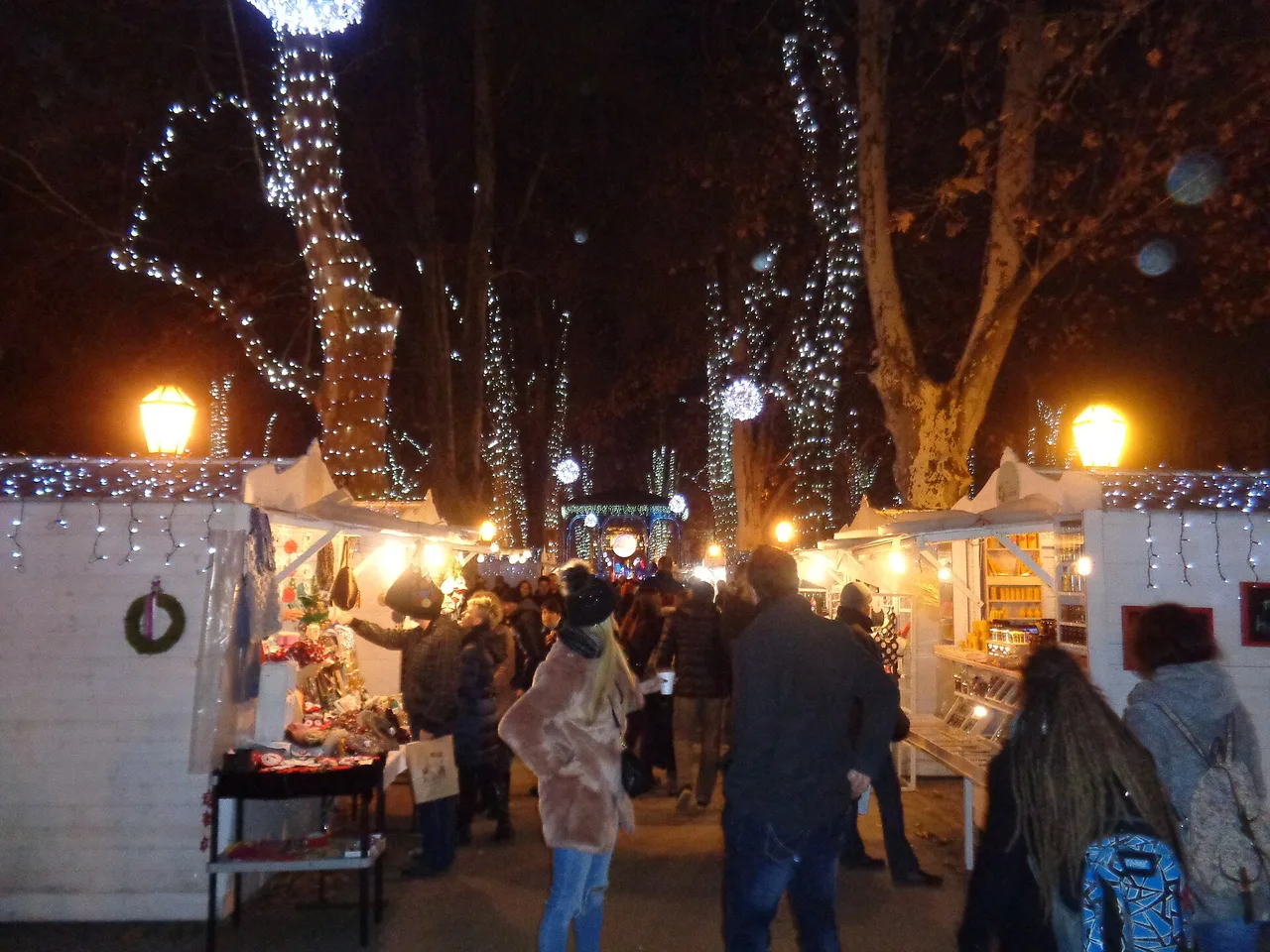 Wooden Advent huts selling crafts under plane trees strung with white lights in a Zagreb park at night