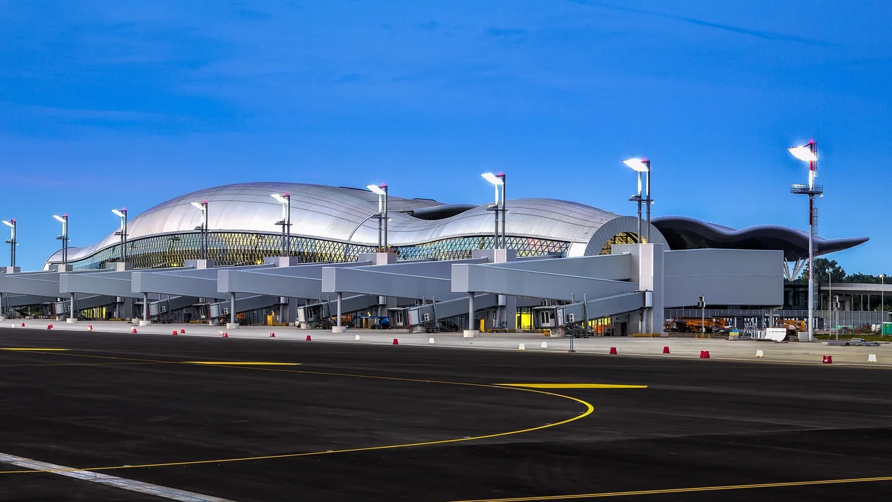 The wave-shaped roof and glass front of Zagreb Airport's passenger terminal at blue hour, seen from the apron with jet bridges and floodlight masts