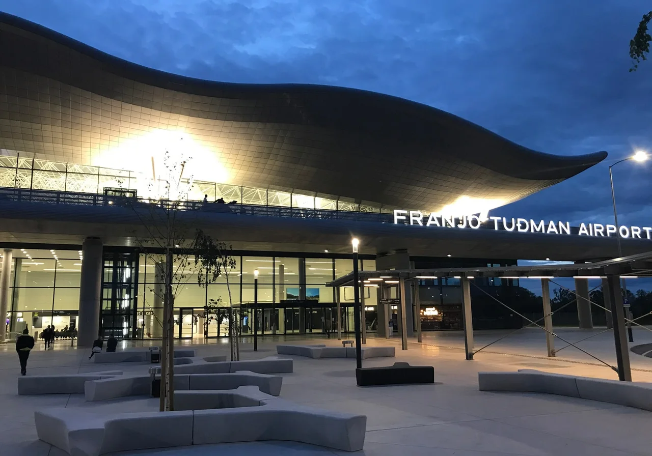 The curved roof and glass front of Franjo Tuđman Airport at dusk, with concrete benches and young trees on the forecourt