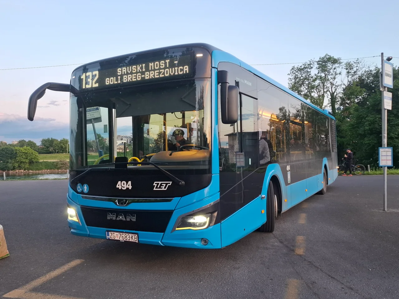 A blue ZET city bus, a MAN Lion's City on line 132, standing at a terminus stop with a river embankment behind it at dusk