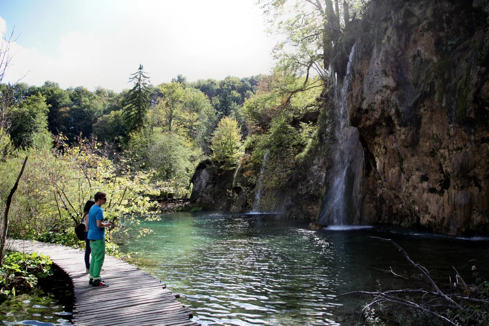 Two visitors on a wooden boardwalk beside a clear green pool and small waterfalls at Plitvice
